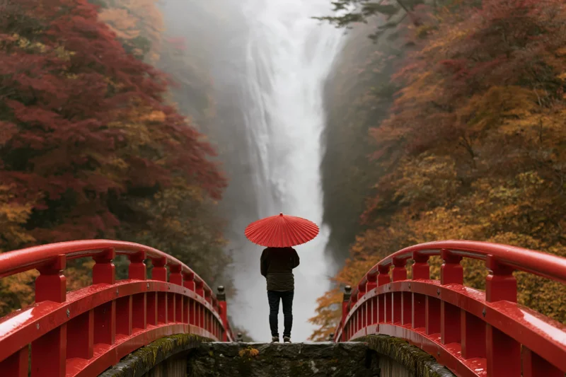 Person with red umbrella standing on red arched bridge facing waterfall surrounded by autumn foliage