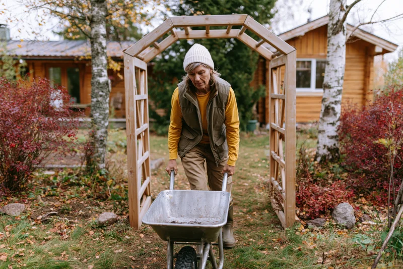 Middle-aged woman pushing metal wheelbarrow through garden archway in autumn yard