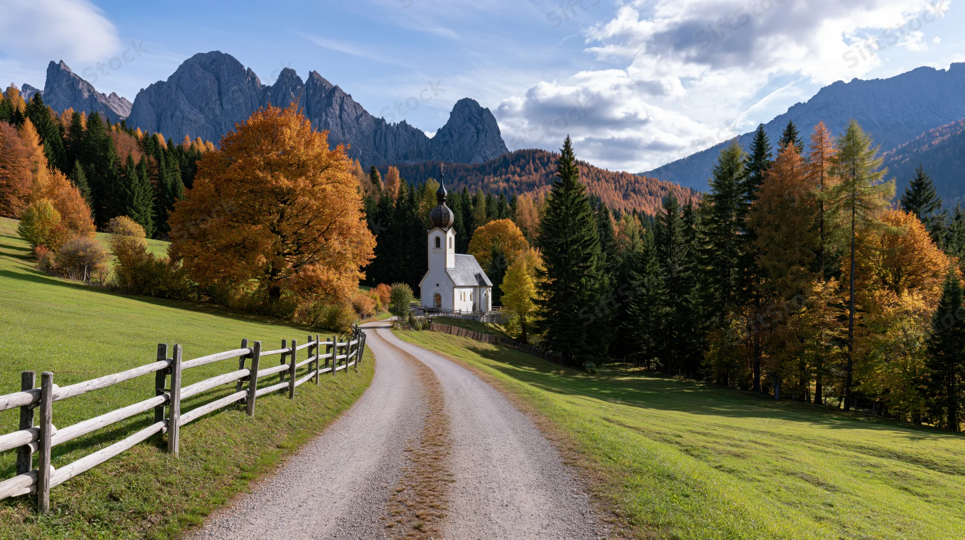 Winding gravel road leading to white alpine church surrounded by autumn trees and mountains