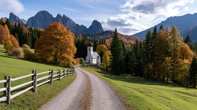 Winding gravel road leading to white alpine church surrounded by autumn trees and mountains