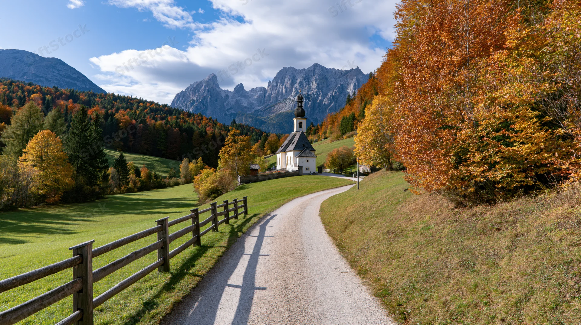 Narrow gravel road curving toward white alpine chapel surrounded by autumn trees and mountains