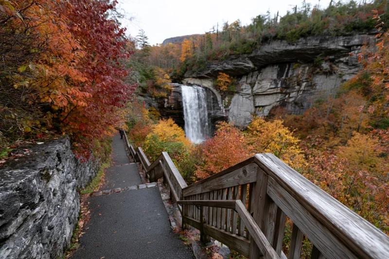 Paved path with wooden railings descending toward a waterfall surrounded by autumn foliage in a forested mountain area