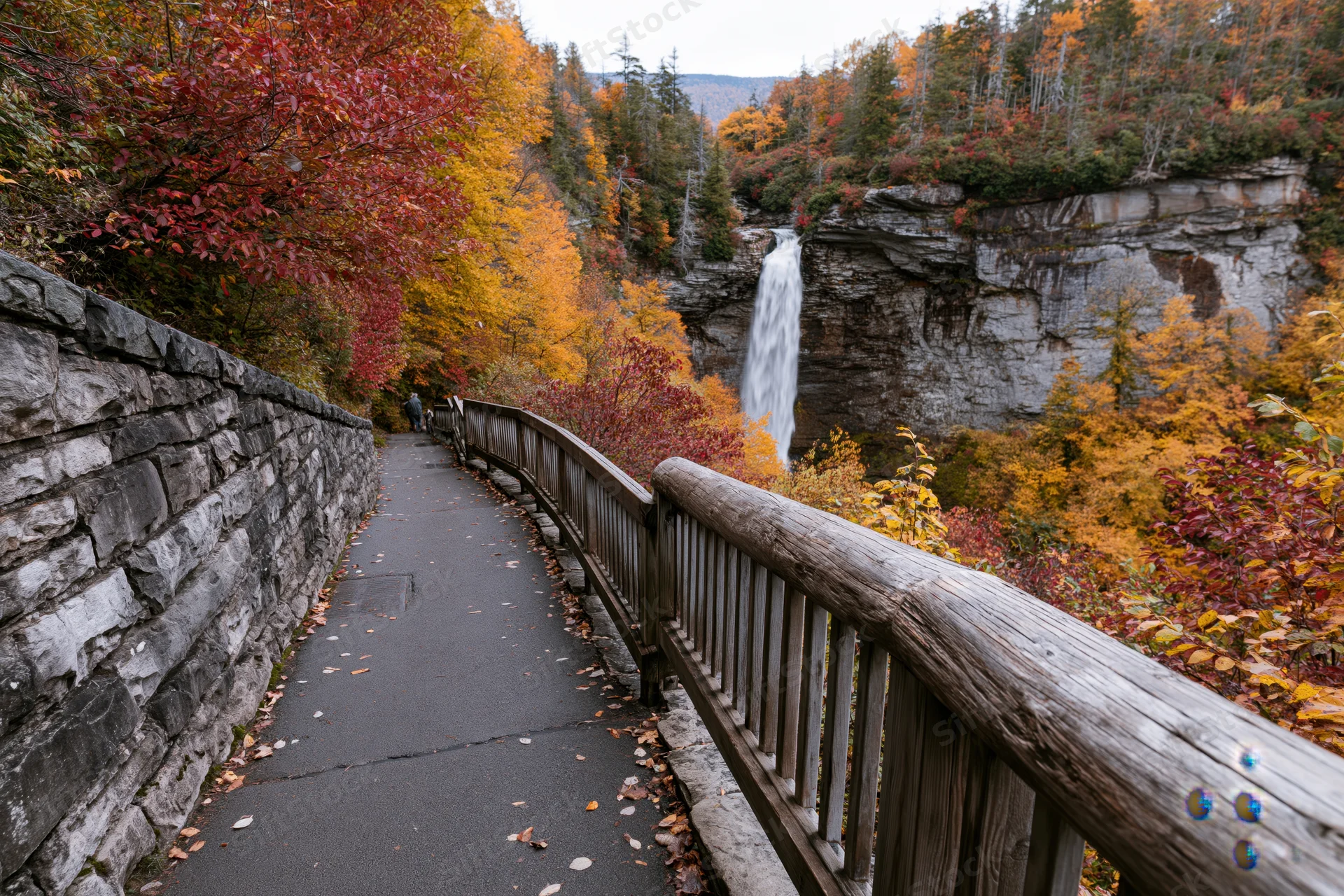 Paved path with wooden railing winding through autumn forest toward waterfall