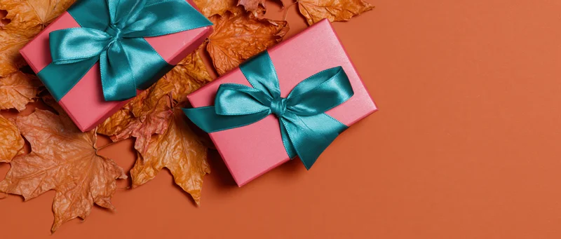 Two pink gift boxes with teal satin bows on dried maple leaves and orange surface