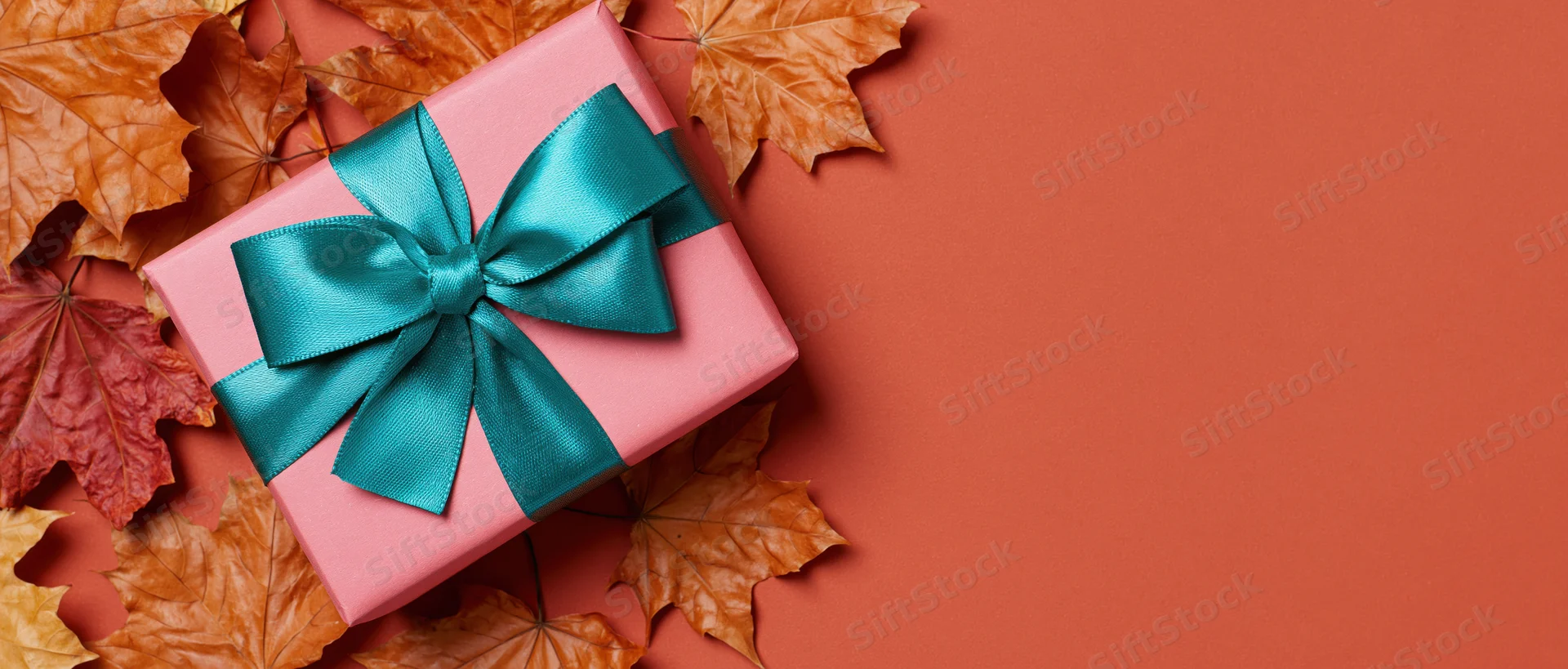 Pink gift box with teal satin bow surrounded by autumn maple leaves on orange surface