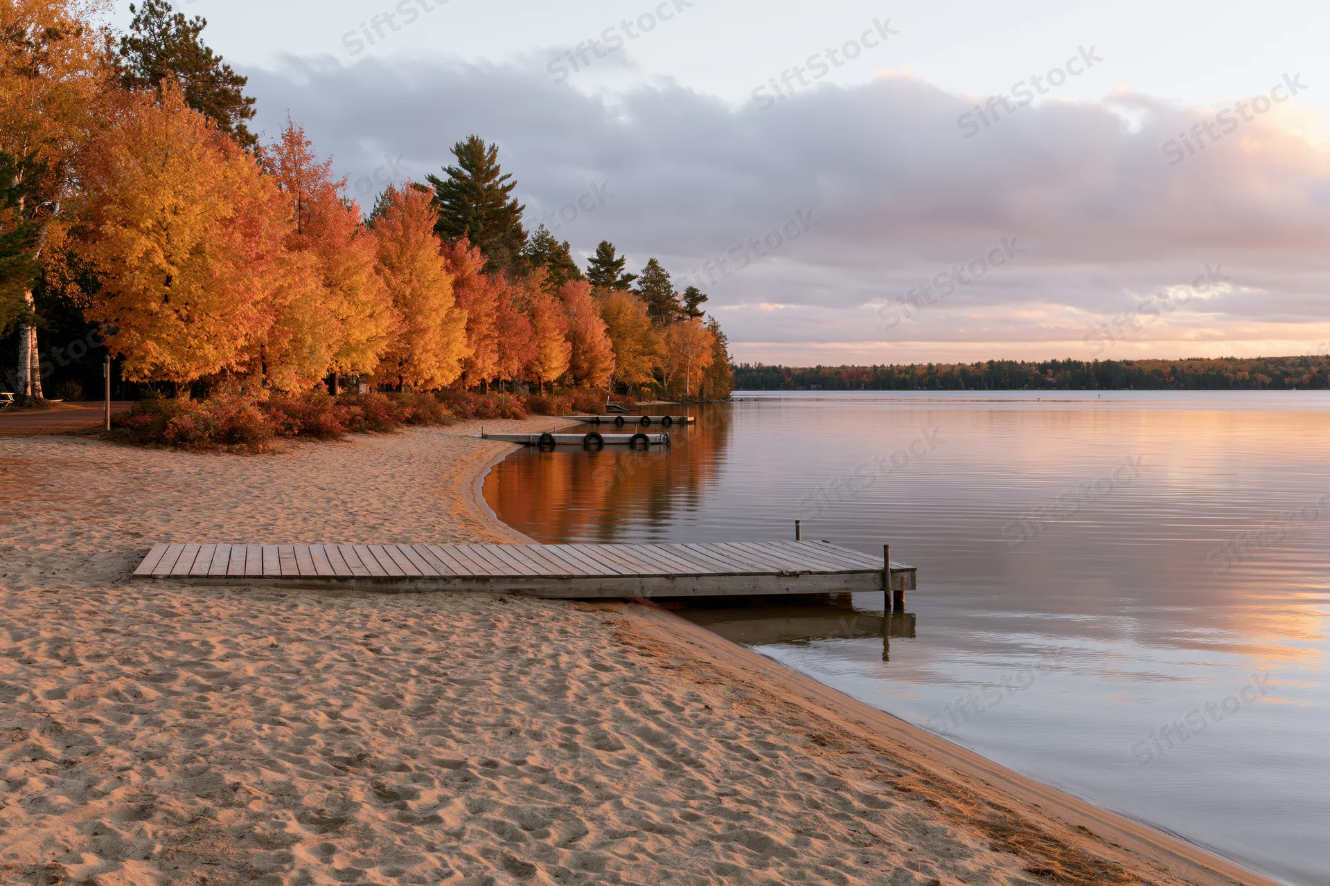 Quiet lakeside beach in autumn with wooden dock extending into calm water beside colorful trees