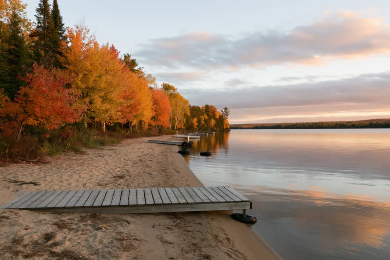 Wooden dock extending from sandy beach into calm lake surrounded by autumn trees at sunset