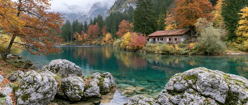 Rustic wooden cabin beside clear turquoise mountain lake surrounded by autumn forest and mossy rocks
