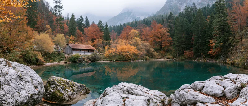 Rustic wooden cabin beside clear turquoise mountain lake surrounded by autumn forest with rock formations in foreground