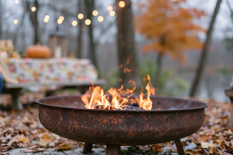 Rusty metal fire pit burning with orange and yellow flames in a leaf-covered backyard during autumn evening