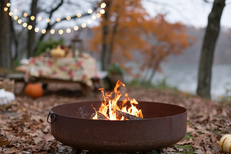 Rusty metal fire pit burning with orange and yellow flames in an autumn outdoor setting near a lake
