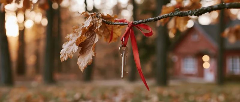 Silver key tied with red ribbon to oak branch in autumn forest with house in background