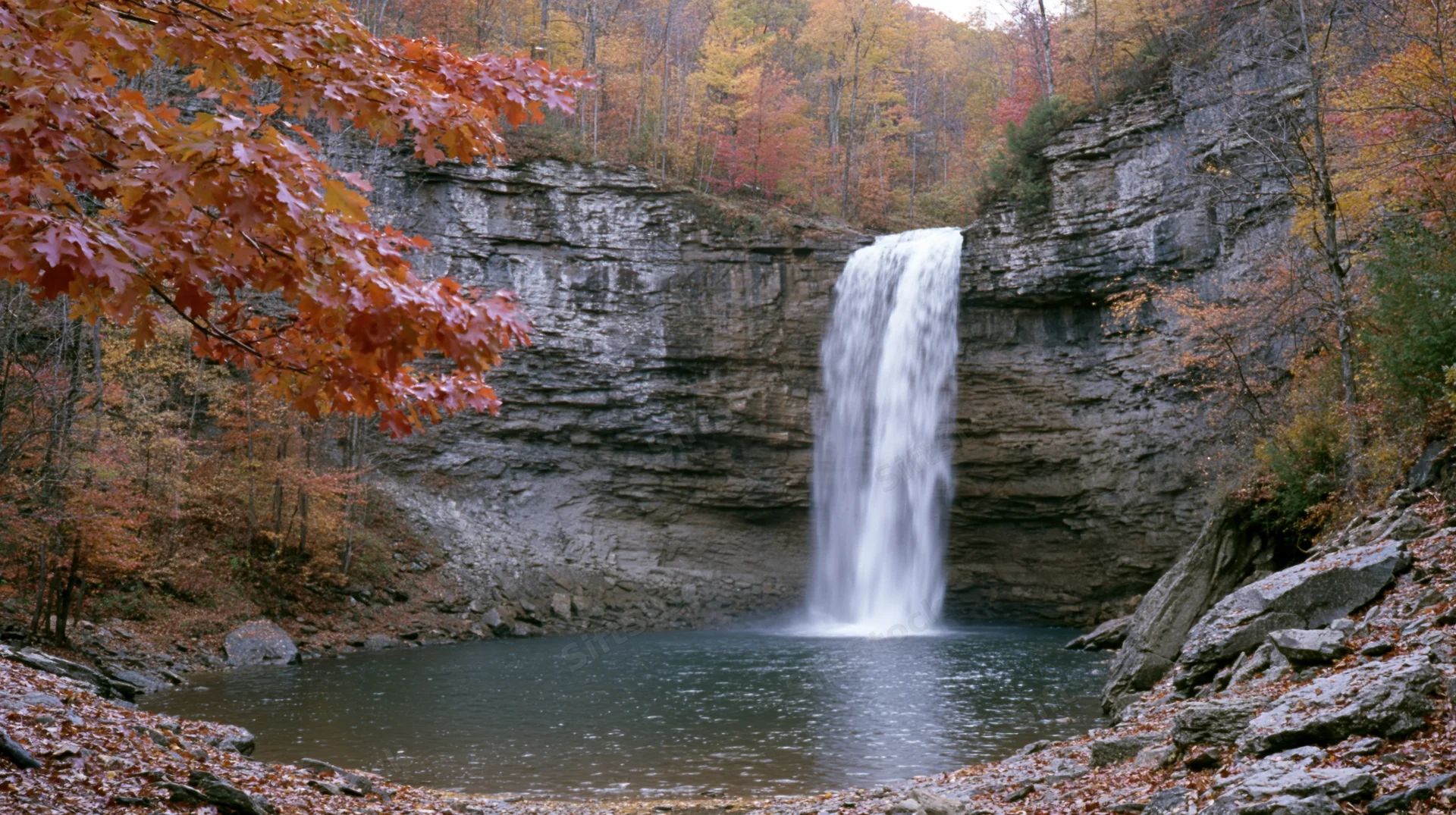 Tall waterfall cascading over layered rock cliff into pool surrounded by autumn forest