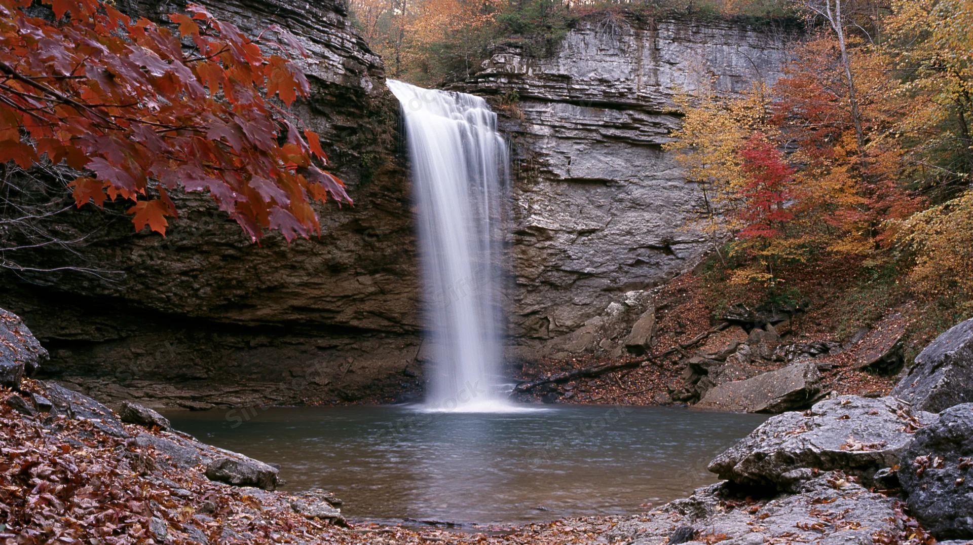Tall waterfall cascading over layered rock cliff into pool surrounded by autumn foliage