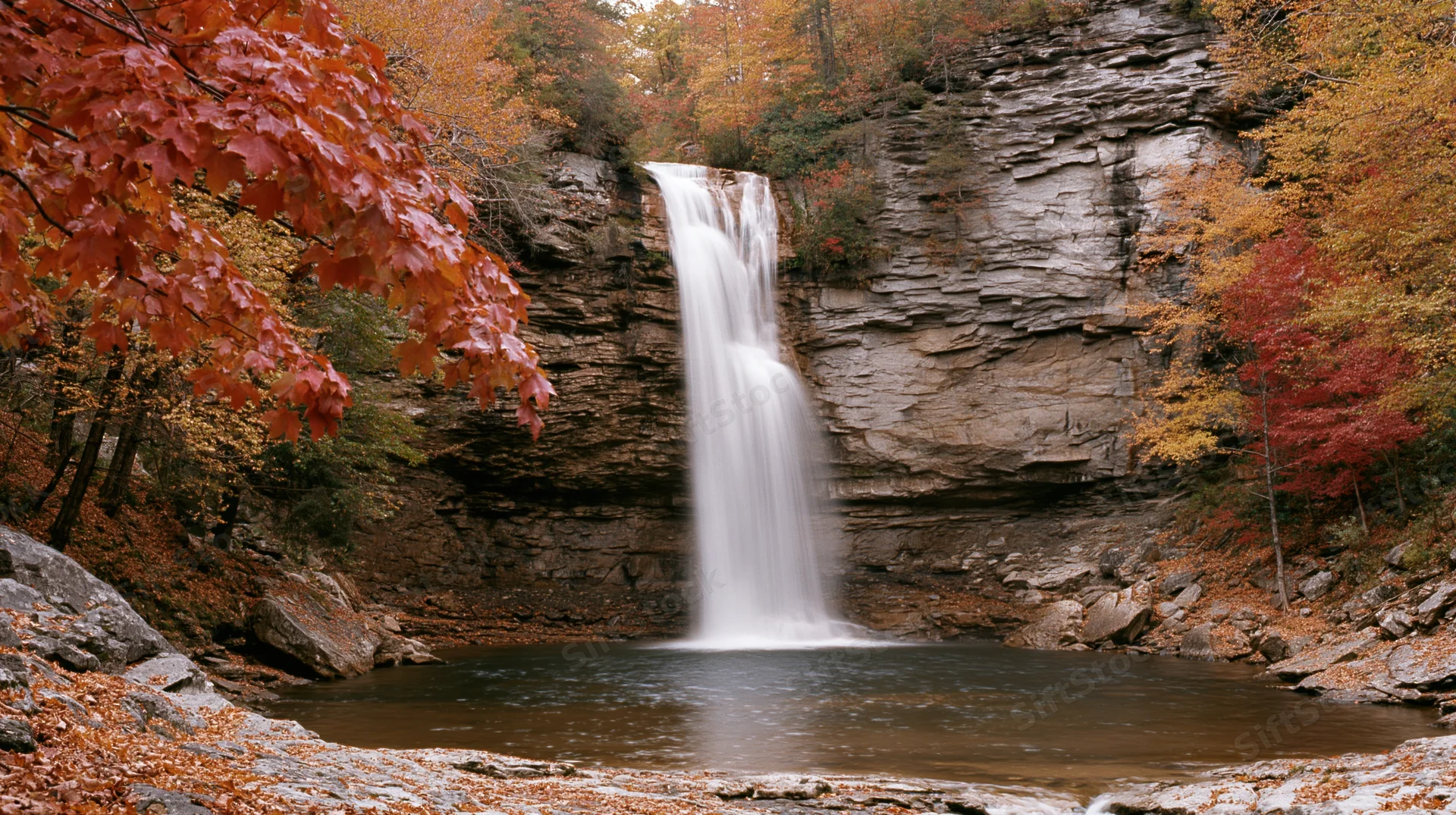Tall waterfall cascading over layered rock cliff surrounded by autumn forest with colorful foliage and stream below