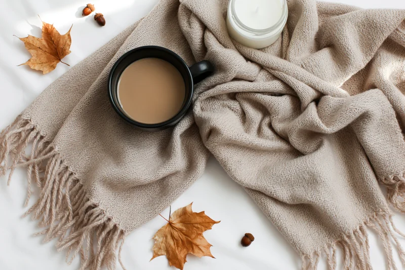 Flat lay of a cozy autumn scene with a knitted scarf, coffee mug, candle, and fallen leaves on a white surface