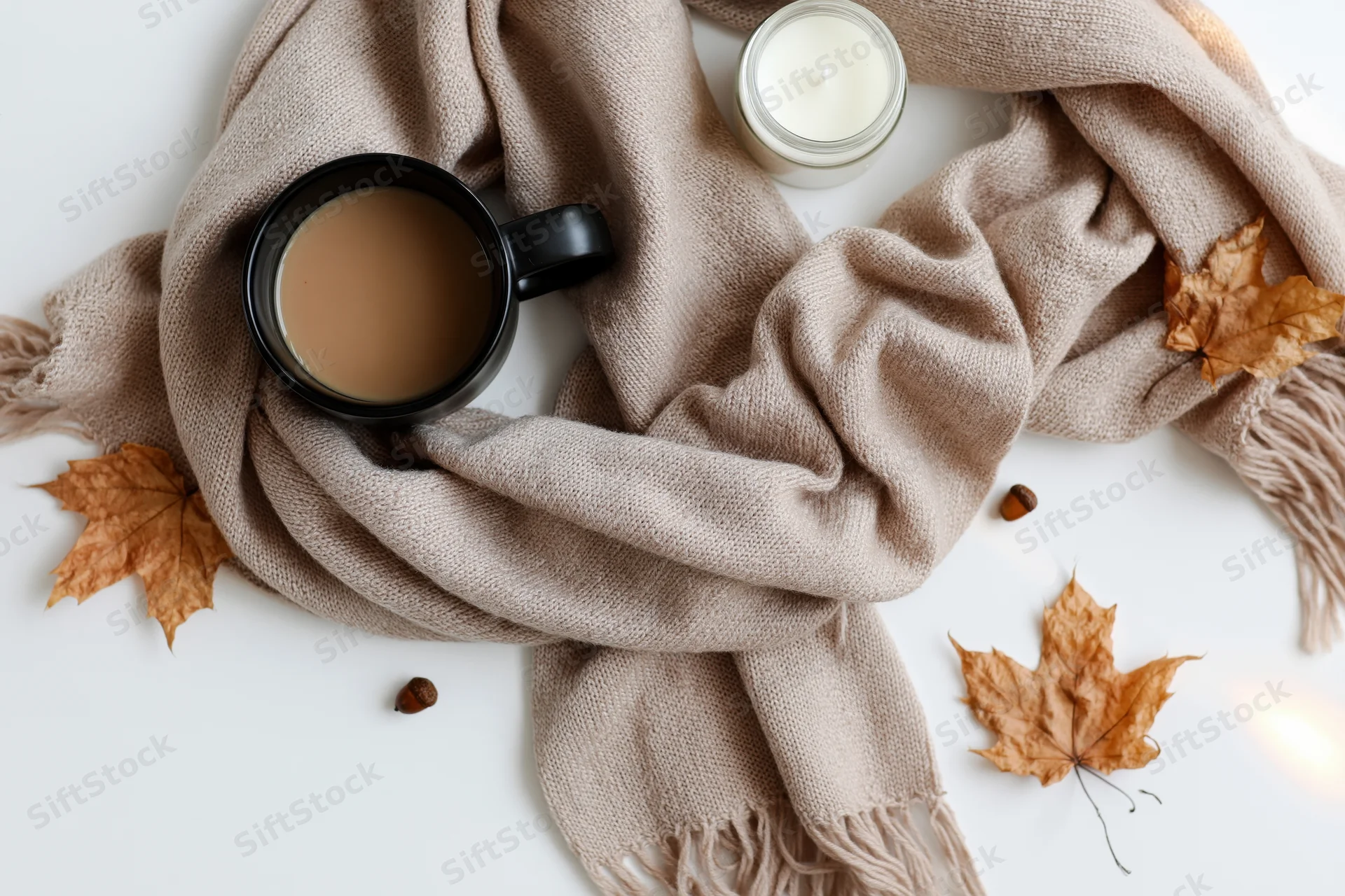 Flat lay of a beige scarf with coffee mug, candle, and autumn leaves on white surface