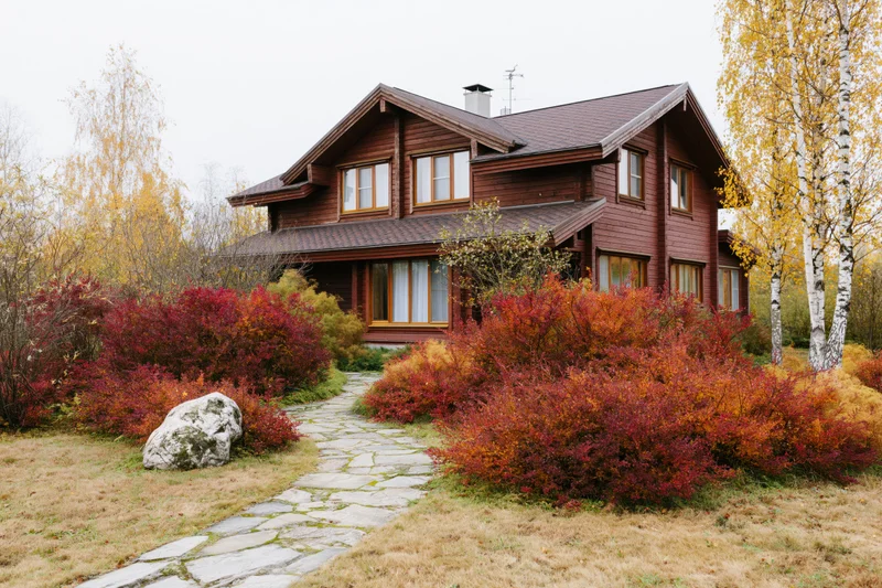 Two-story brown wooden house with dark shingle roof surrounded by autumn foliage and stone pathway