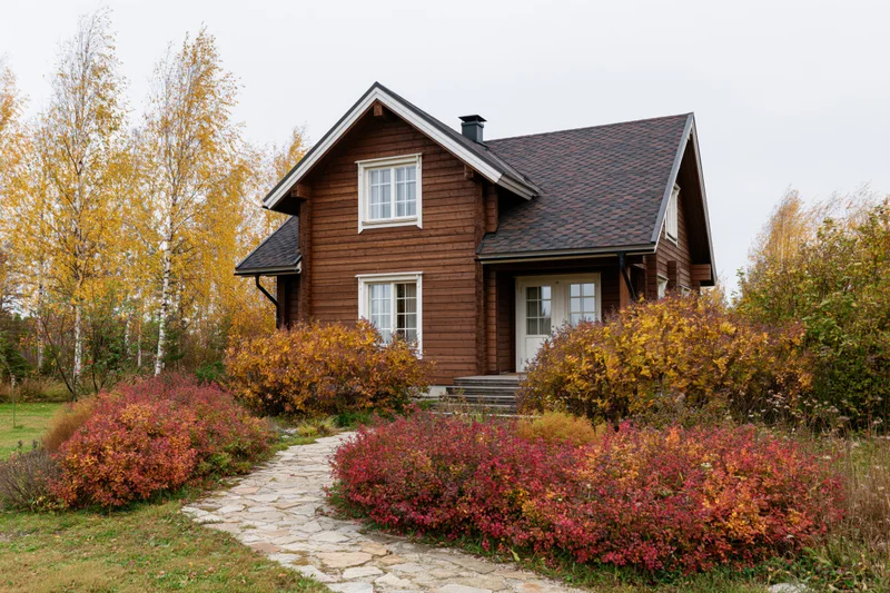Brown wooden two-story country house with dark shingle roof surrounded by autumn foliage and stone pathway