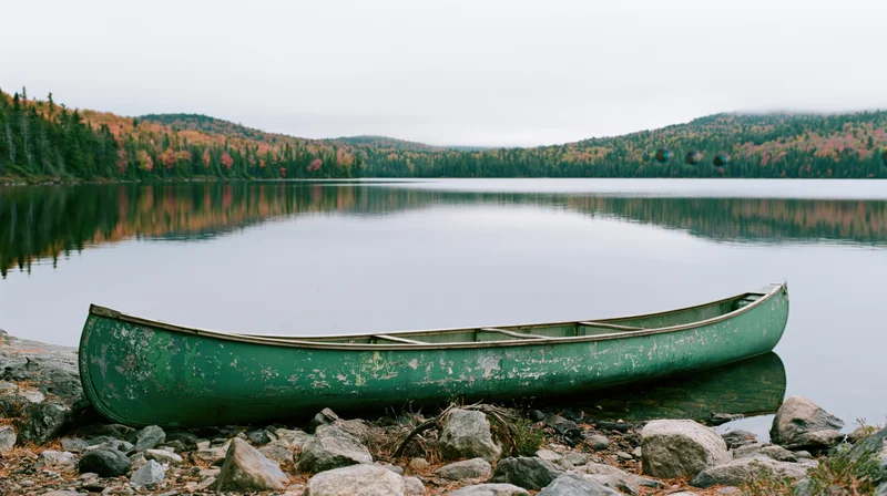 Weathered green canoe resting on rocky shoreline beside calm lake with autumn forest reflection