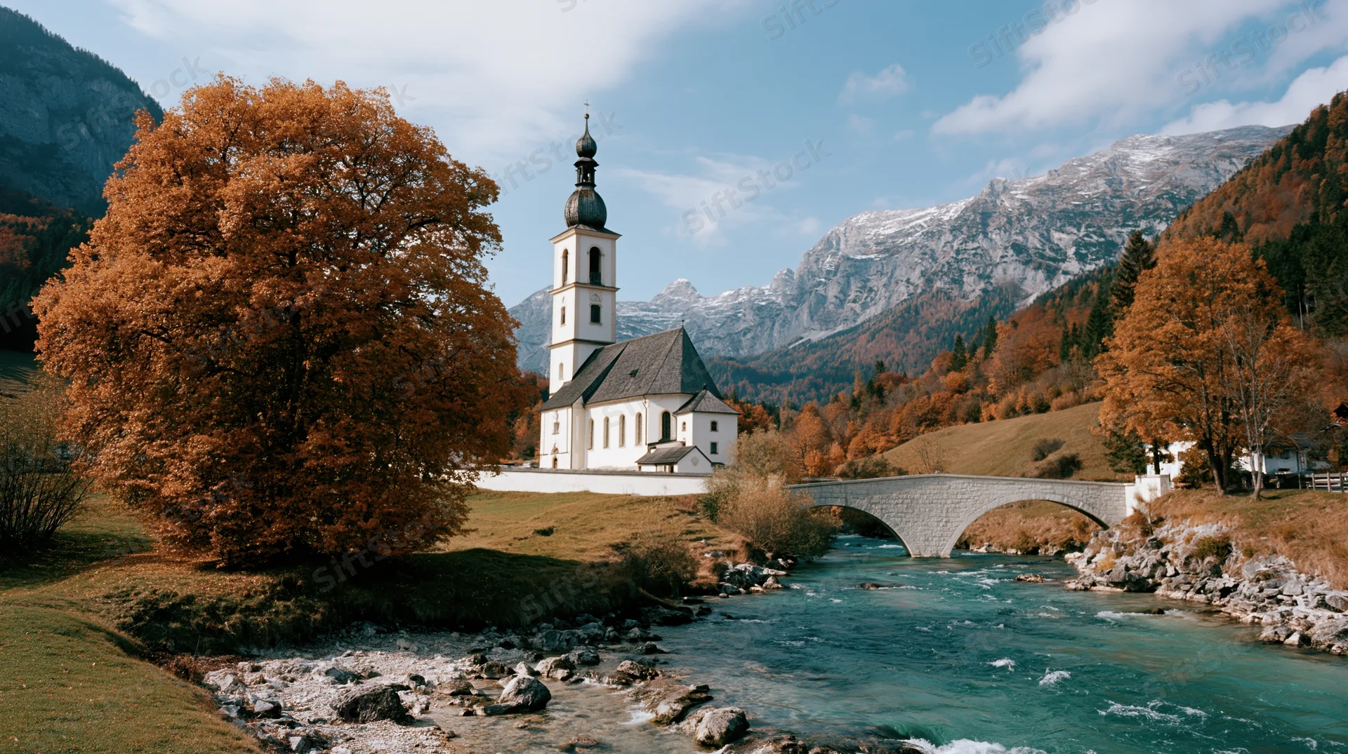 White bavarian church with square steeple beside a river and stone bridge in alpine valley during autumn