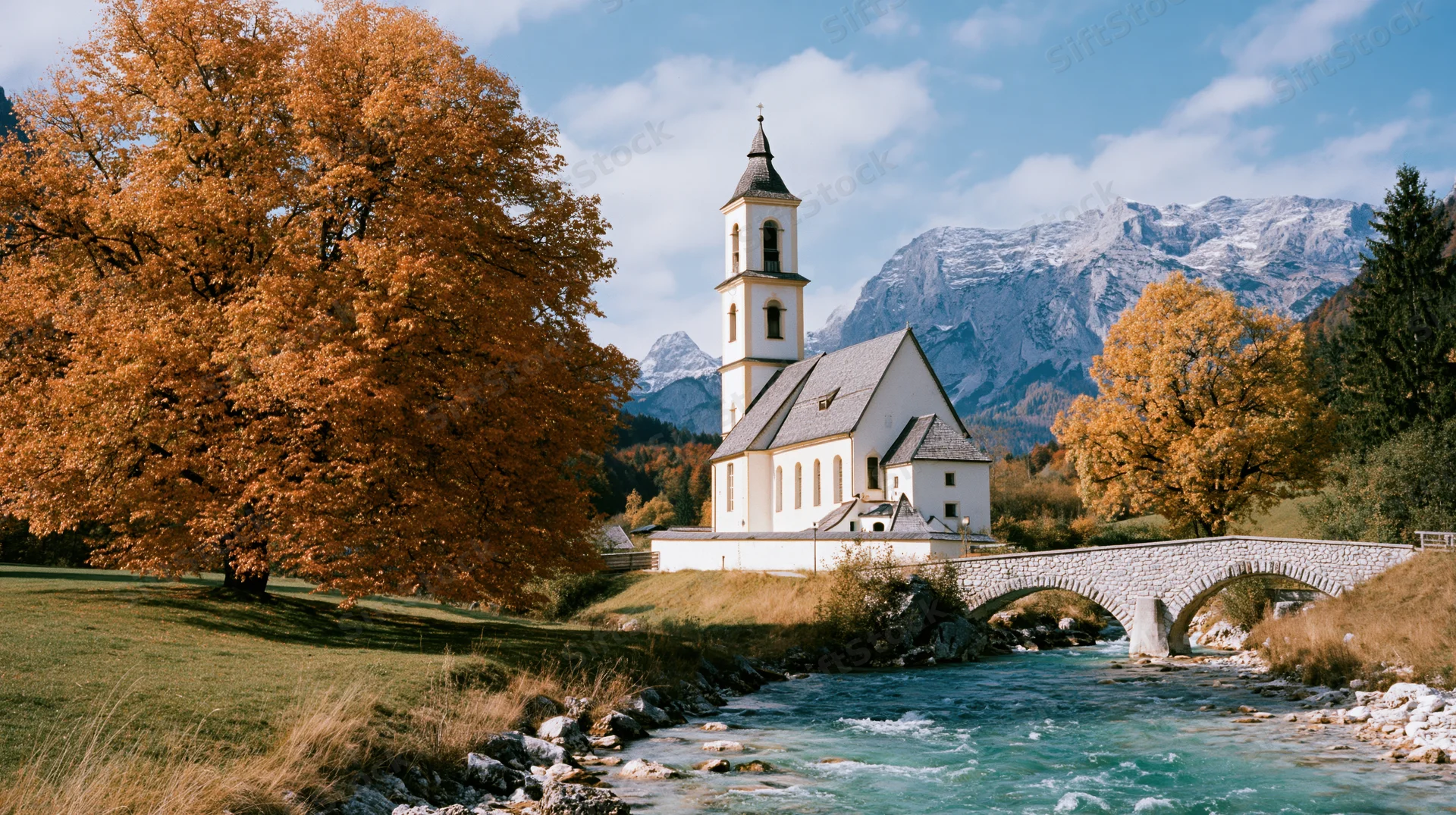 White bavarian parish church with square steeple beside a river and stone bridge in autumn alpine landscape