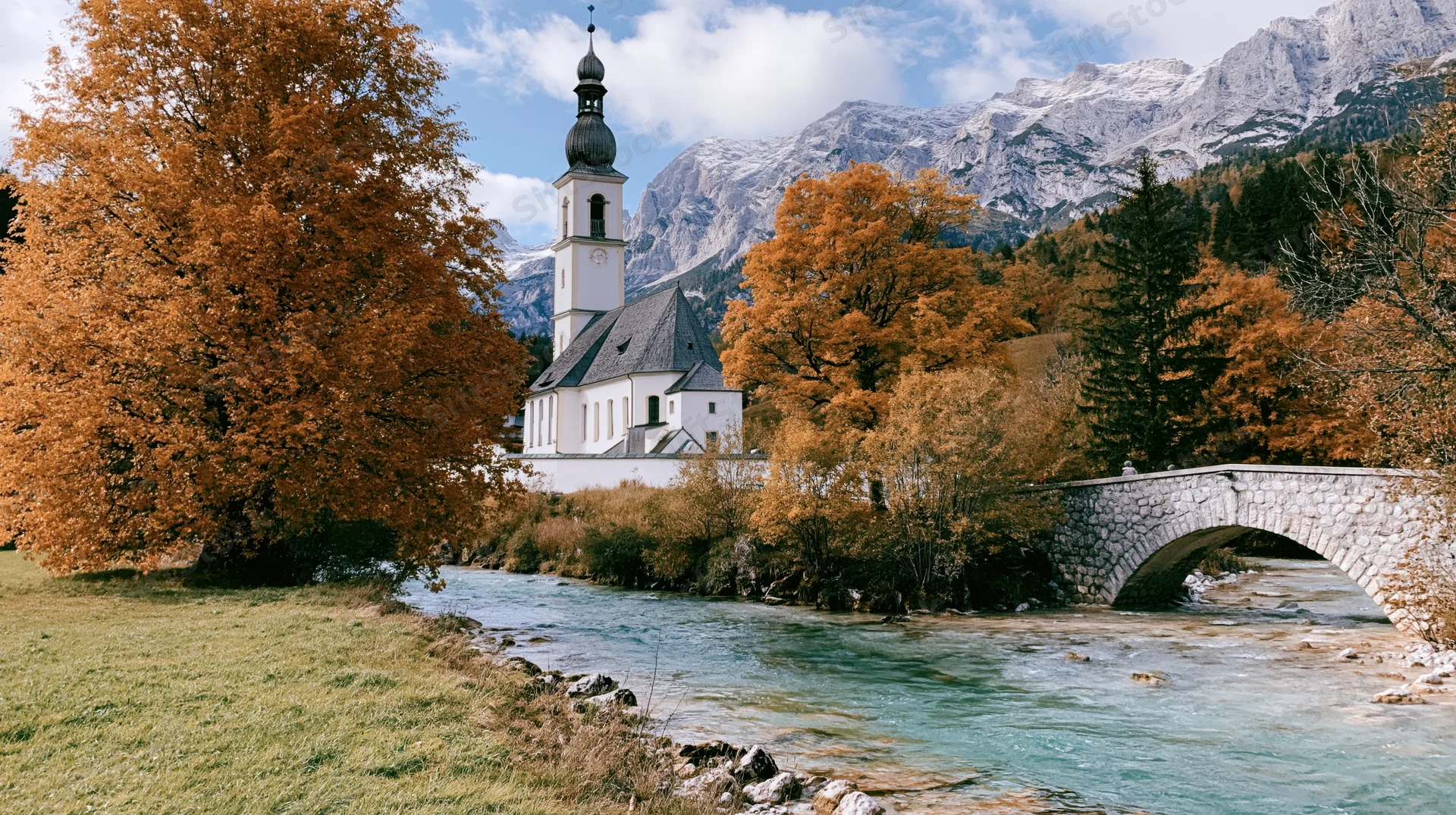 White bavarian parish church with square steeple beside a river and stone bridge in autumnal alpine landscape