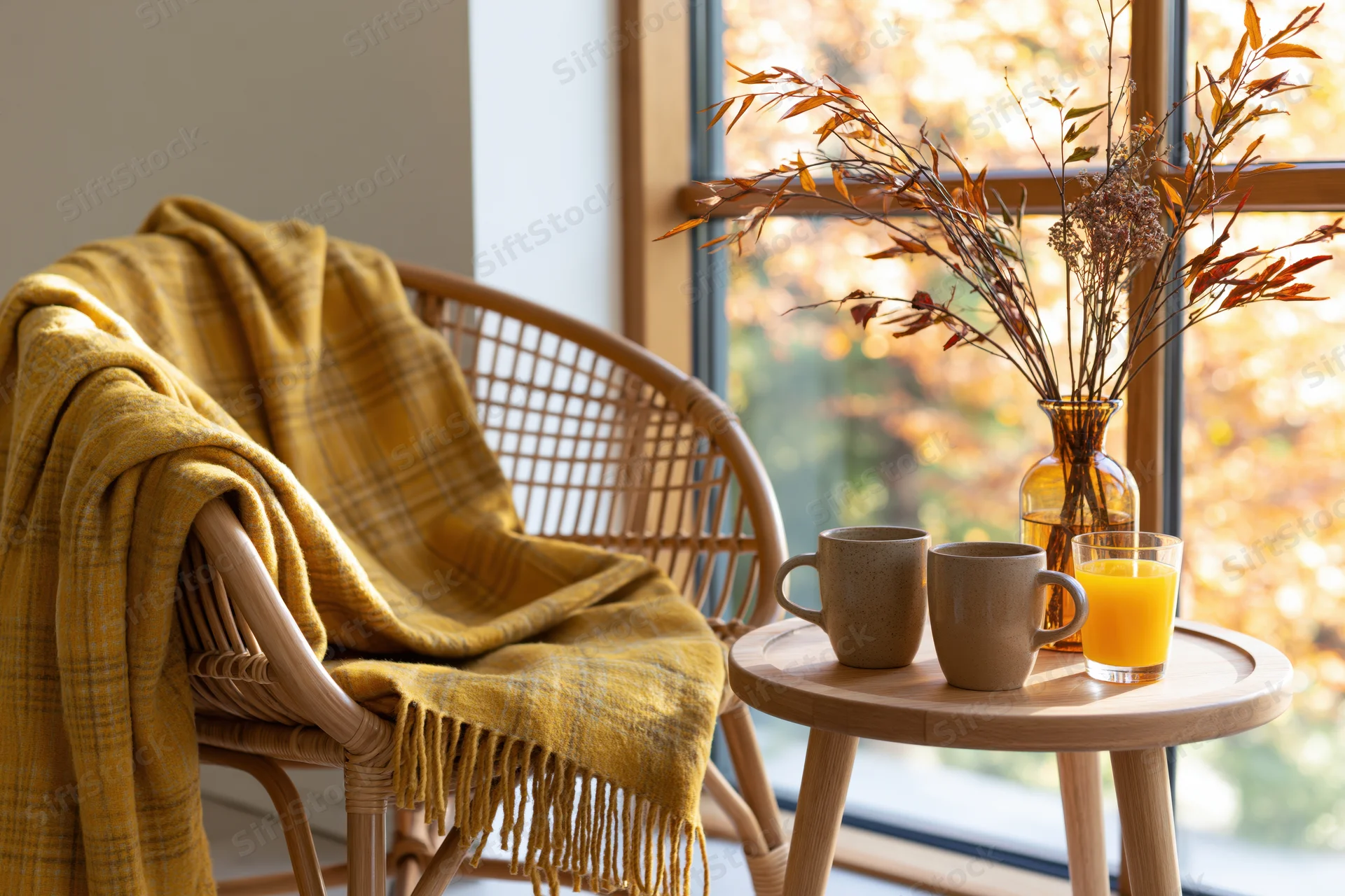 Wicker chair with yellow plaid blanket beside a wooden table holding mugs and orange juice near a window with autumn foliage view