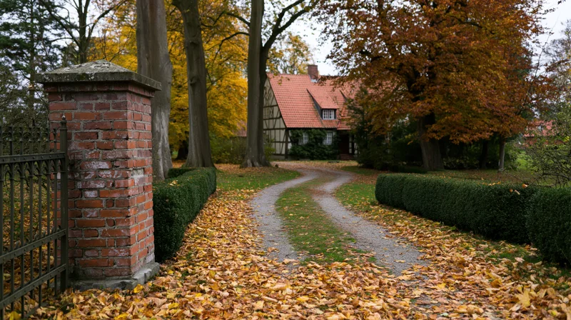 Winding gravel path covered in fallen autumn leaves leading to a half-timbered house surrounded by trees and hedges