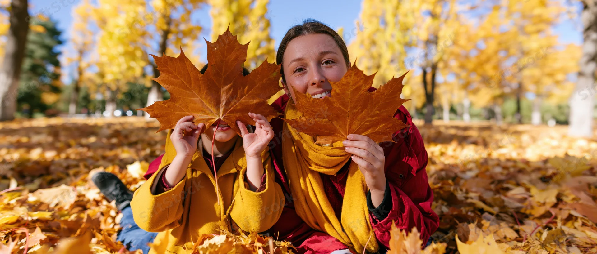 Woman and child sitting in autumn leaves in a park, holding maple leaves over their faces, smiling