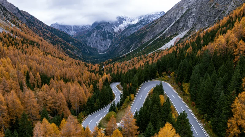 Winding alpine road through autumn forested valley with snow-capped peaks above