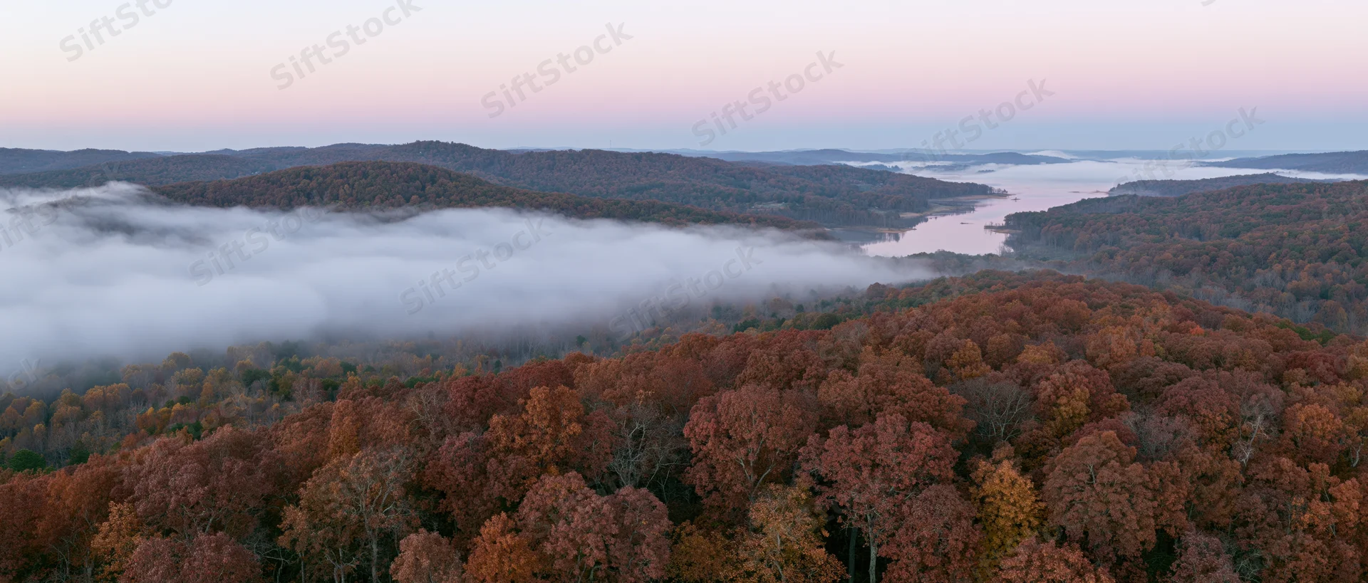 Aerial panoramic view of autumn forest canopy with misty river and mountains at dawn