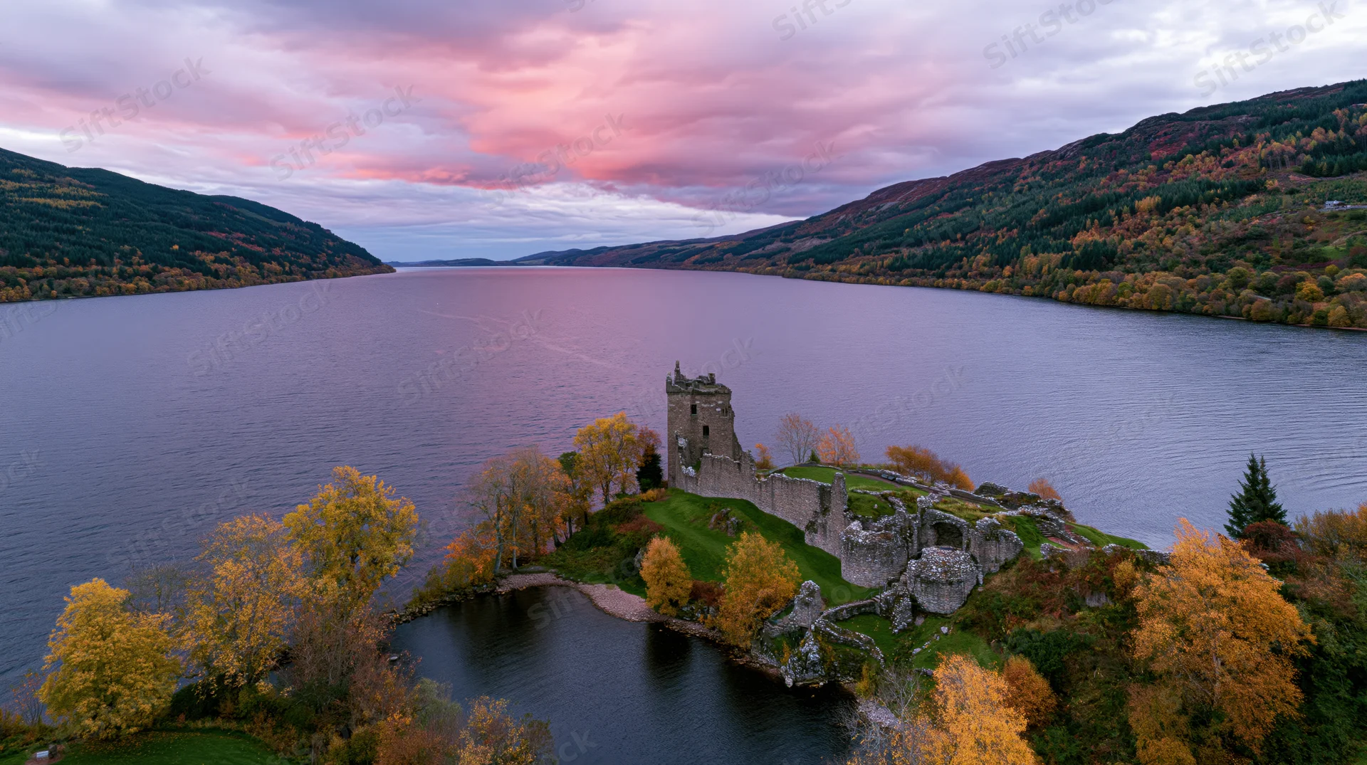 Aerial view of urquhart castle ruins on a rocky promontory overlooking loch ness surrounded by autumn trees and mountains