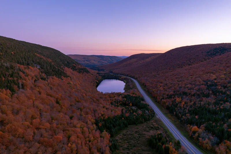 Aerial view of a winding road through a mountain valley covered in autumn foliage with a lake at dusk