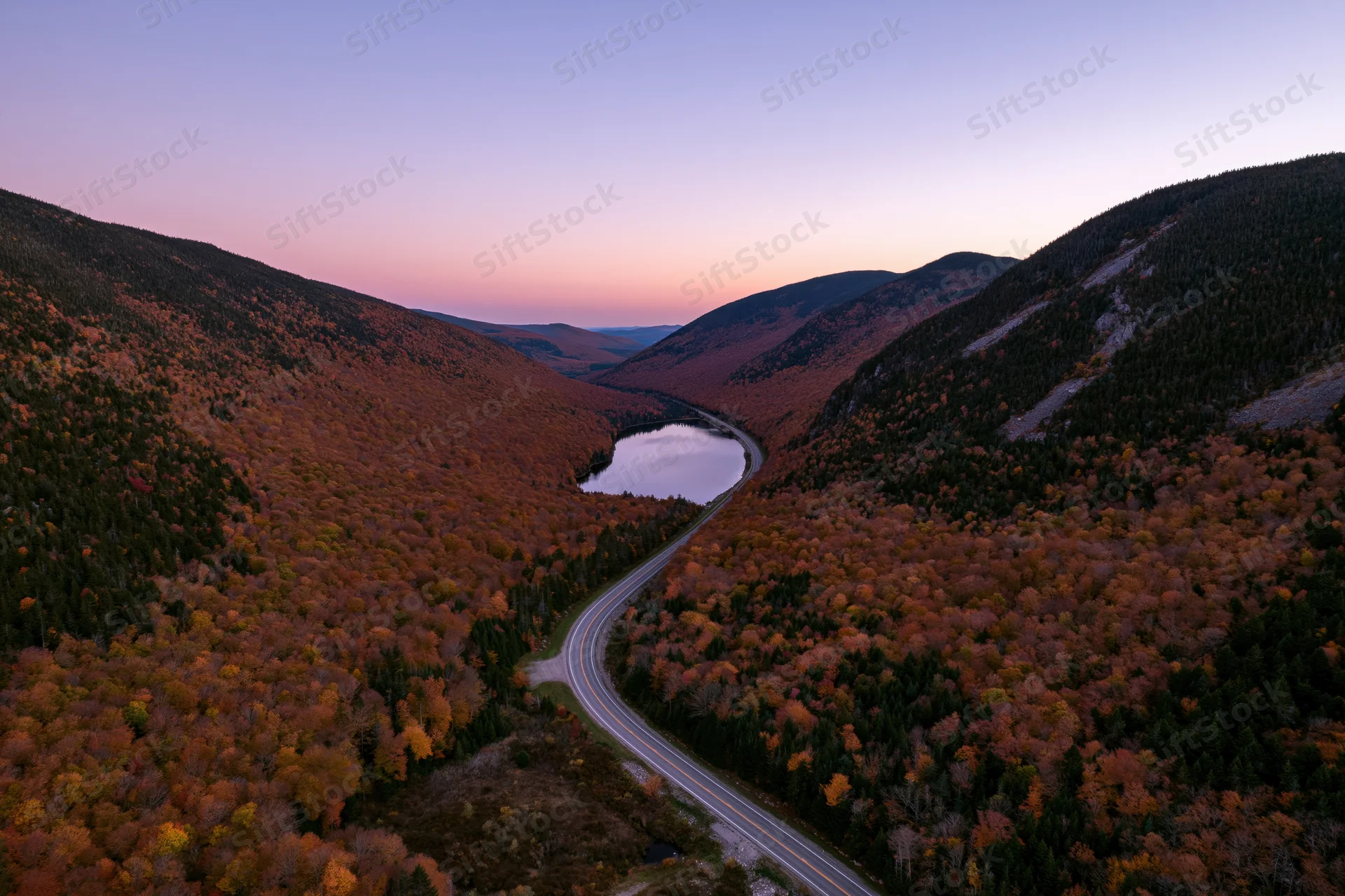 Aerial view of a winding road through a mountain valley covered in autumn foliage at sunset