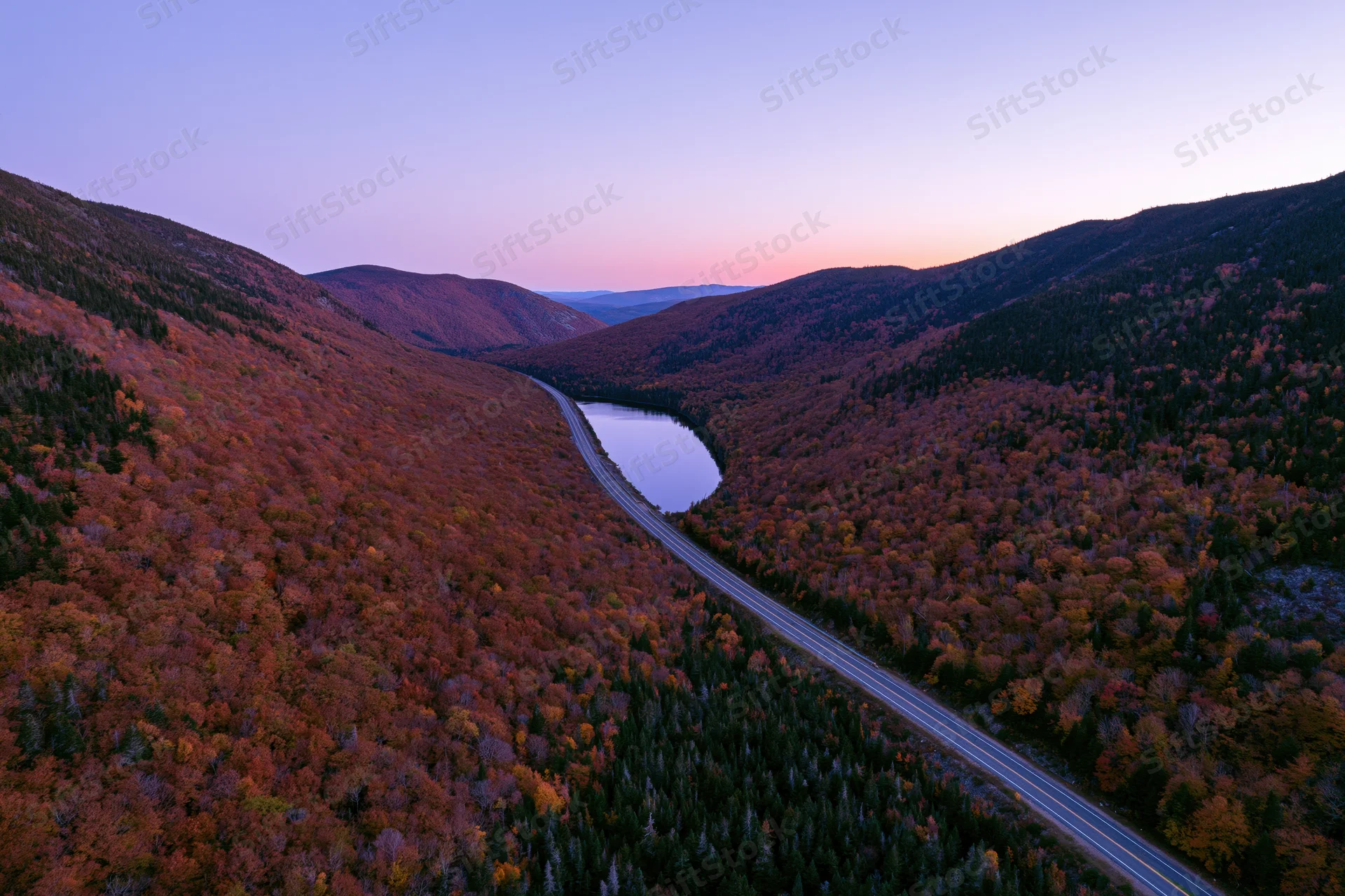 Aerial view of a mountain valley with autumn foliage and a winding road alongside a lake at dusk