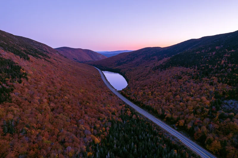 Aerial view of a mountain valley with autumn foliage and a winding road alongside a lake at dusk