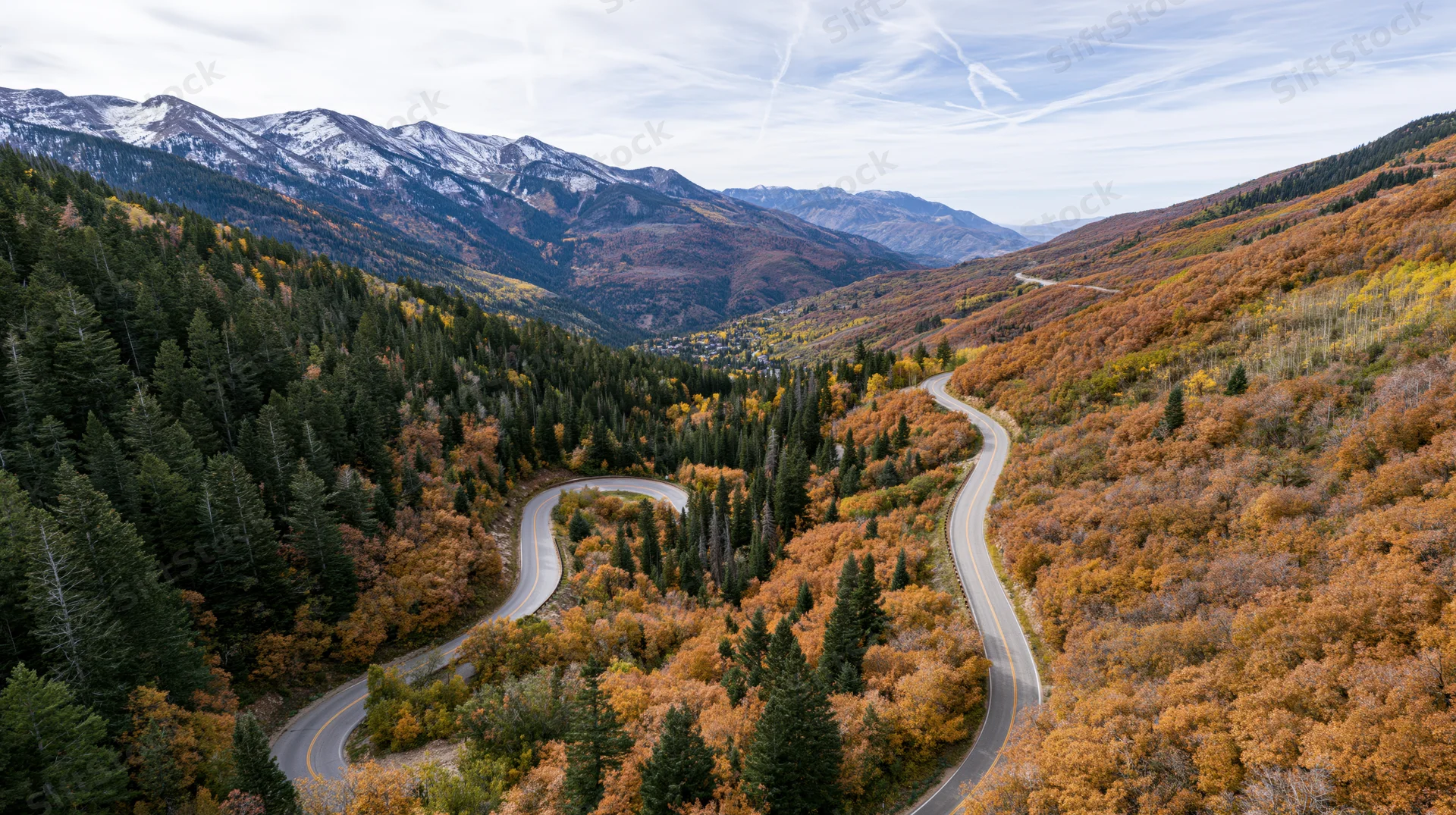 Winding alpine road through autumn forested valley with snow-capped mountains in background