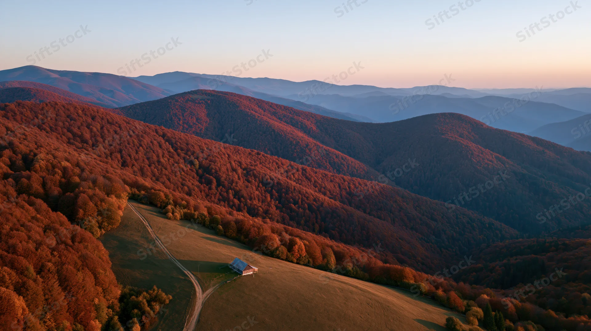 Aerial view of rolling carpathian hills covered in autumn forest with a barn on a meadow at sunset