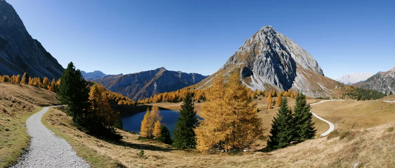 Winding gravel path through alpine valley with autumn trees and mountain lake under clear blue sky