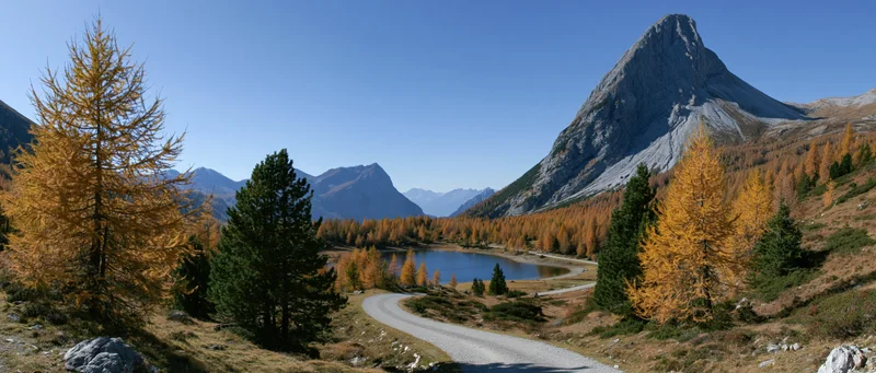 Winding gravel path through alpine valley with autumn trees and mountain peak under clear blue sky