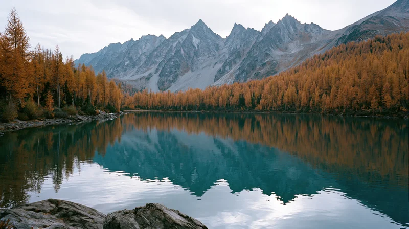 Alpine lake reflecting jagged mountains and golden autumn trees under cloudy sky