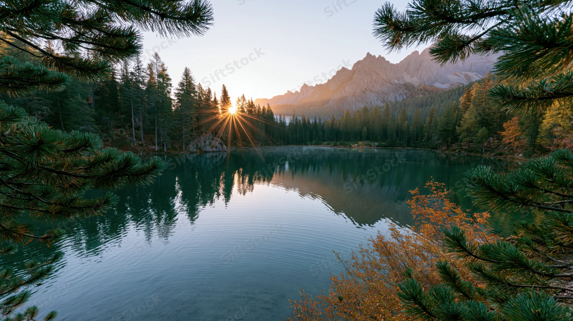 Alpine lake at sunrise with still water reflecting mountains and forest through pine branches