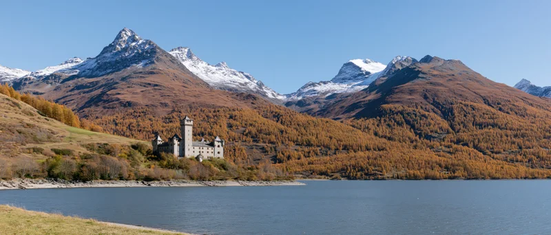 Alpine lake shoreline with castle nestled among autumn-colored hills and snow-capped mountains under clear blue sky