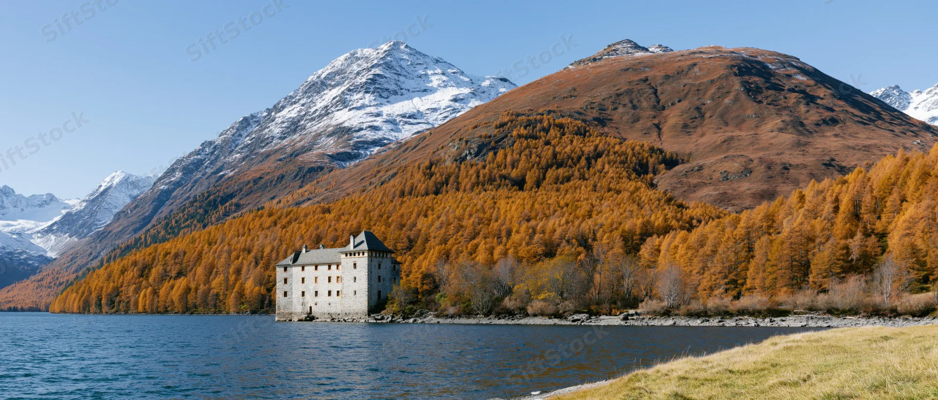 Wide panoramic view of alpine lake shoreline with autumn forest and snow-capped mountains