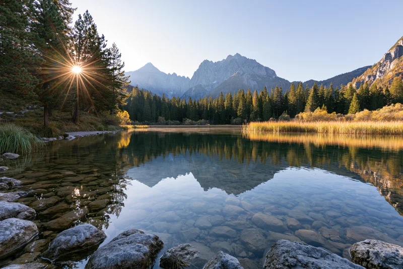 Alpine lake with clear shallow water reflecting pine trees and mountains at sunrise
