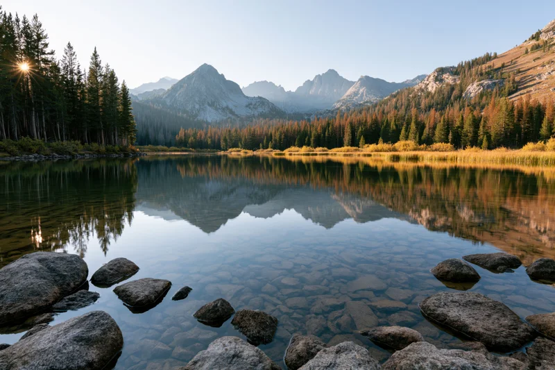 Alpine lake with clear shallow water reflecting pine trees and mountains at sunrise