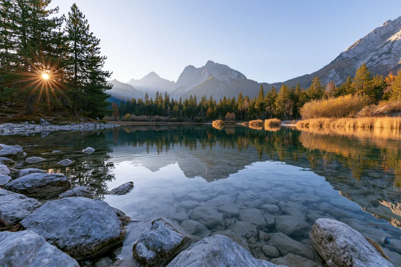Alpine lake with clear shallow water reflecting pine trees and mountains at sunrise