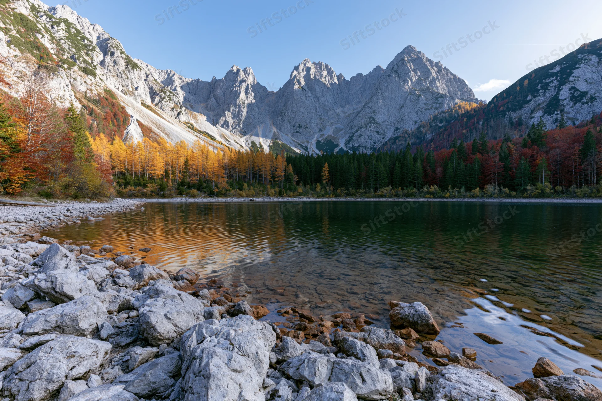 Alpine lake surrounded by jagged limestone peaks and autumn-colored forest under clear blue sky