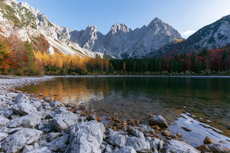 Alpine lake surrounded by jagged limestone peaks and autumn-colored forest under clear blue sky